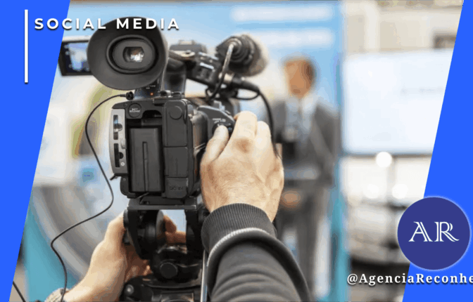 Câmera de vídeo profissional em primeiro plano sendo operada durante uma entrevista coletiva, com um jornalista desfocado ao fundo em um ambiente de estúdio ou evento.