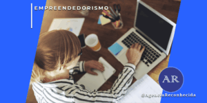 Mulher loira trabalhando em mesa de escritório com notebook, cadernos e relatórios, vista superior, com o selo da Agência Reconhecida no canto inferior.