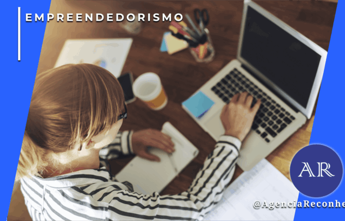 Mulher loira trabalhando em mesa de escritório com notebook, cadernos e relatórios, vista superior, com o selo da Agência Reconhecida no canto inferior.