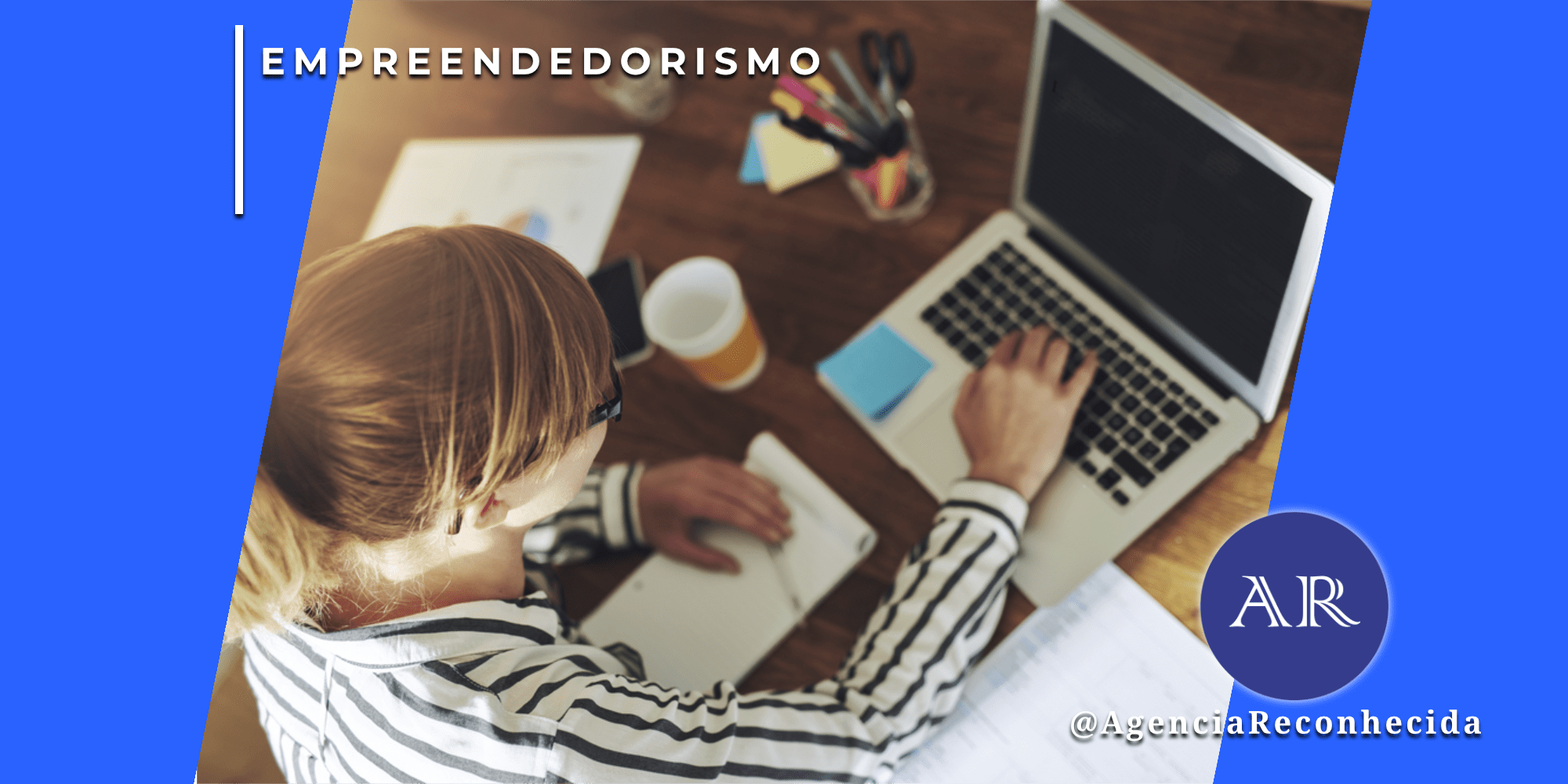 Mulher loira trabalhando em mesa de escritório com notebook, cadernos e relatórios, vista superior, com o selo da Agência Reconhecida no canto inferior.