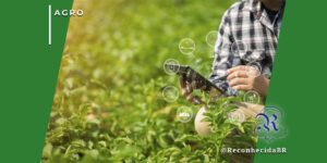Close-up de um profissional do agronegócio segurando um tablet em uma plantação verde sob a luz do sol. Ícones digitais brancos sobrepostos representam tecnologia agro: conectividade, energia solar, sensores e sustentabilidade. No canto superior esquerdo está o selo 'AGRO' e no inferior direito o logotipo do portal Reconhecida com o @ReconhecidaBR.