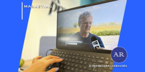 Visão em primeira pessoa de uma mão feminina com unhas lilases digitando em um teclado de notebook. Na tela do computador, um vídeo mostra uma entrevista jornalística com um homem de óculos (Walmir Rampinelli), identificado por um GC e um microfone da emissora NSC. No canto superior esquerdo, o selo 'MARKETING' em fundo azul e, no inferior direito, o logotipo AR da Agência Reconhecida com o @AgenciaReconhecida.