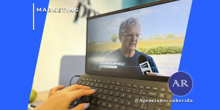 Visão em primeira pessoa de uma mão feminina com unhas lilases digitando em um teclado de notebook. Na tela do computador, um vídeo mostra uma entrevista jornalística com um homem de óculos (Walmir Rampinelli), identificado por um GC e um microfone da emissora NSC. No canto superior esquerdo, o selo 'MARKETING' em fundo azul e, no inferior direito, o logotipo AR da Agência Reconhecida com o @AgenciaReconhecida.