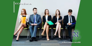 Cinco candidatos a emprego sentados em uma sala de espera, vestindo trajes formais e segurando pastas coloridas. Eles expressam descontentamento, tédio e ansiedade. No canto superior esquerdo, o selo 'ECONOMIA' e, no inferior direito, o logotipo da Reconhecida com o @ReconhecidaBR.