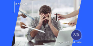 Um homem sentado à mesa com as mãos no rosto, expressando estresse e sobrecarga. Ao seu redor, várias mãos estendem tablets, documentos e smartphones em sua direção. No canto superior esquerdo, o selo 'GESTÃO' em fundo azul e, no inferior direito, o logotipo AR da Agência Reconhecida com o @AgenciaReconhecida.
