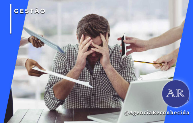 Um homem sentado à mesa com as mãos no rosto, expressando estresse e sobrecarga. Ao seu redor, várias mãos estendem tablets, documentos e smartphones em sua direção. No canto superior esquerdo, o selo 'GESTÃO' em fundo azul e, no inferior direito, o logotipo AR da Agência Reconhecida com o @AgenciaReconhecida.