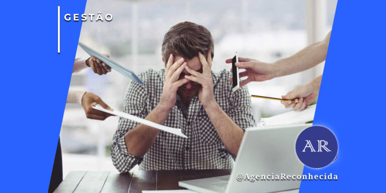 Um homem sentado à mesa com as mãos no rosto, expressando estresse e sobrecarga. Ao seu redor, várias mãos estendem tablets, documentos e smartphones em sua direção. No canto superior esquerdo, o selo 'GESTÃO' em fundo azul e, no inferior direito, o logotipo AR da Agência Reconhecida com o @AgenciaReconhecida.