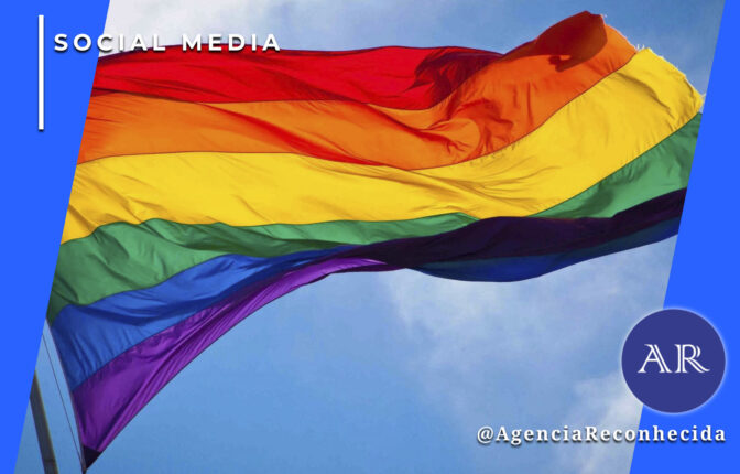 Uma grande bandeira com as cores do arco-íris (símbolo LGBT+) hasteada e ondulando contra um céu azul com nuvens claras. No canto superior esquerdo, o selo 'SOCIAL MEDIA' sobre fundo azul e, no inferior direito, o logotipo AR da Agência Reconhecida com o @AgenciaReconhecida.