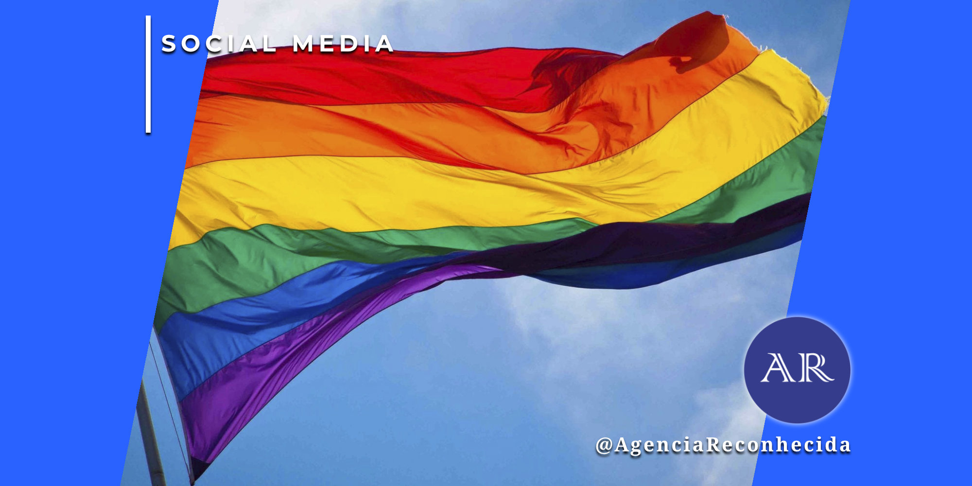 Uma grande bandeira com as cores do arco-íris (símbolo LGBT+) hasteada e ondulando contra um céu azul com nuvens claras. No canto superior esquerdo, o selo 'SOCIAL MEDIA' sobre fundo azul e, no inferior direito, o logotipo AR da Agência Reconhecida com o @AgenciaReconhecida.