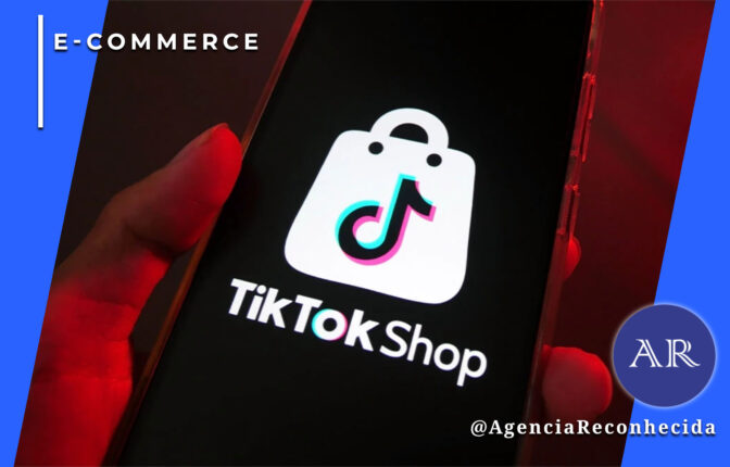 Mão segurando um smartphone que exibe o logotipo branco do TikTok Shop em uma tela preta. O logotipo consiste em uma sacola de compras com o símbolo musical icônico do TikTok no centro. No canto superior esquerdo, o selo 'E-COMMERCE' sobre fundo azul e, no inferior direito, o logotipo AR da Agência Reconhecida com o @AgenciaReconhecida.