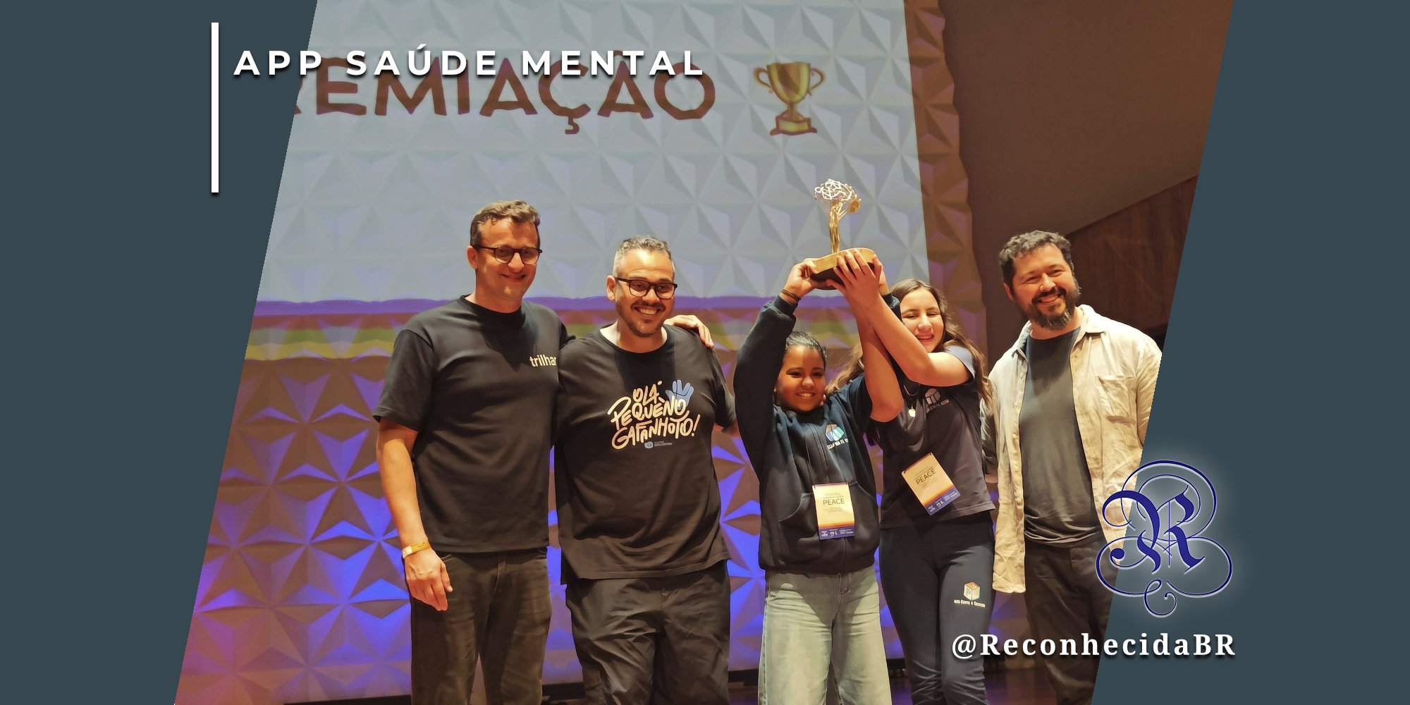 Foto de premiação em um palco com três homens adultos e duas jovens estudantes sorridentes. As alunas seguram juntas um troféu dourado em formato de árvore. Ao fundo, um telão exibe a palavra 'PREMIAÇÃO' e um ícone de troféu. No canto superior esquerdo, o selo 'APP SAÚDE MENTAL' em fundo branco e, no inferior direito, o logotipo do portal Reconhecida com o @ReconhecidaBR.