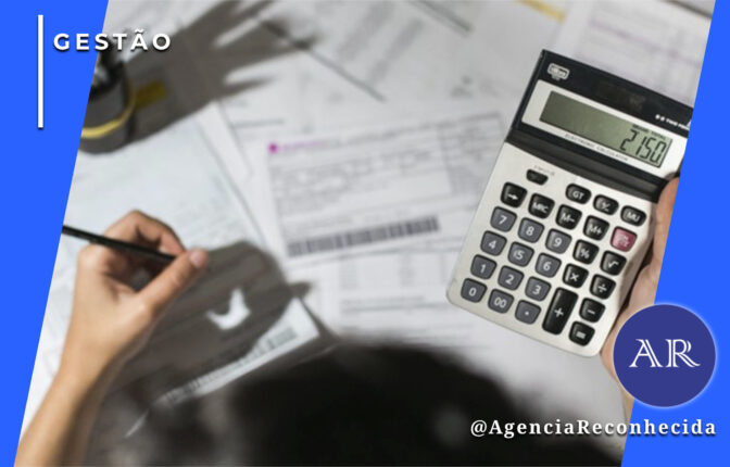 Visão aérea de uma mesa de escritório com diversos boletos e documentos financeiros espalhados. Uma pessoa segura uma calculadora eletrônica exibindo o número 2150, enquanto outra mão escreve com um lápis em um papel. No canto superior esquerdo, o selo 'GESTÃO' em fundo azul e, no inferior direito, o logotipo AR da Agência Reconhecida com o @AgenciaReconhecida.