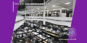 Visão aérea panorâmica do pavilhão da Bienal em São Paulo ocupado pelo Salão do Artesanato Raízes Brasileiras. Diversos estandes pretos e brancos organizados em níveis diferentes exibem artesanatos regionais sob iluminação clara. No canto superior esquerdo, o selo 'SALÃO DO ARTESANATO' em branco sobre fundo roxo e, no inferior direito, o logotipo do portal Reconhecida com o @ReconhecidaBR.