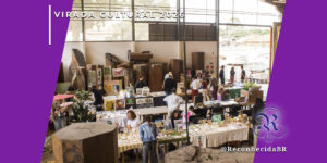 Visão ampla do interior de um galpão industrial com pé-direito alto, onde ocorre a Feira da Gamela durante a Virada Cultural 2026. Diversas bancadas exibem produtos artesanais, gravuras e objetos de decoração, cercadas por grandes troncos de madeira esculpidos. Pessoas circulam e interagem com os expositores. No topo esquerdo, o selo 'VIRADA CULTURAL 2026' em branco e, no inferior direito, o logotipo do portal Reconhecida com o @ReconhecidaBR.
