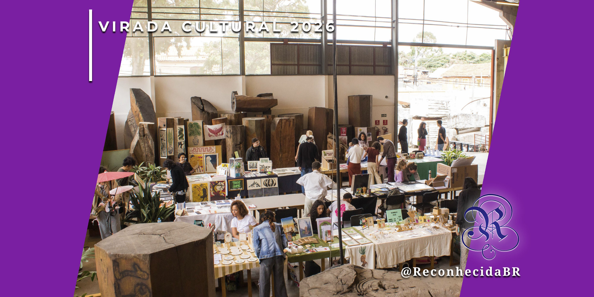 Visão ampla do interior de um galpão industrial com pé-direito alto, onde ocorre a Feira da Gamela durante a Virada Cultural 2026. Diversas bancadas exibem produtos artesanais, gravuras e objetos de decoração, cercadas por grandes troncos de madeira esculpidos. Pessoas circulam e interagem com os expositores. No topo esquerdo, o selo 'VIRADA CULTURAL 2026' em branco e, no inferior direito, o logotipo do portal Reconhecida com o @ReconhecidaBR.