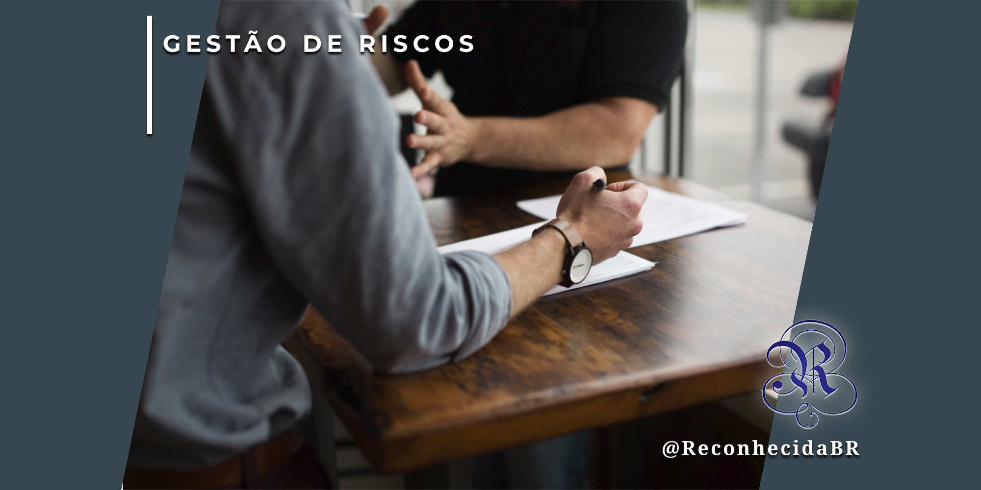 Visão aproximada das mãos de um profissional com relógio de pulso escrevendo em um documento sobre uma mesa de madeira. À frente, outra pessoa gesticula durante uma conversa de consultoria. No canto superior esquerdo, o selo 'GESTÃO DE RISCOS' em fundo branco e, no inferior direito, o logotipo do portal Reconhecida com o @ReconhecidaBR.