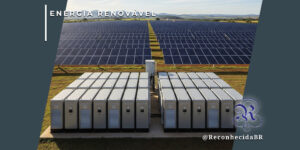 Vista de uma usina solar com extensas fileiras de painéis fotovoltaicos ao fundo. Em primeiro plano, dois conjuntos de grandes baterias industriais cinzas estão instalados sobre uma base de concreto. No canto superior esquerdo, o selo 'ENERGIA RENOVÁVEL' sobre fundo azul; no inferior direito, o logotipo do portal Reconhecida com o @ReconhecidaBR.