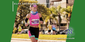 Mulher sorridente correndo em uma maratona ao ar livre, vestindo top rosa, bermuda de compressão preta e rosa, manguitos pretos e óculos de sol espelhados. Ela usa um número de peito da 'Maratona Internacional de Floripa'. No canto superior esquerdo, o selo 'ELAS ESTÃO DOMINANDO' em branco e, no inferior direito, o logotipo do portal Reconhecida com o @ReconhecidaBR.