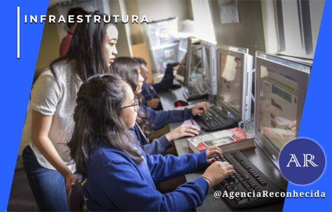 Três jovens estudantes de origem asiática em um laboratório de informática. Uma moça em pé, vestindo camiseta branca, observa atentamente o monitor de outra estudante sentada, que digita em um teclado preto. Ao fundo, uma terceira pessoa também utiliza um computador. No canto superior esquerdo, o selo 'INFRAESTRUTURA' em branco e, no inferior direito, o logotipo da Agência Reconhecida com o @AgenciaReconhecida.