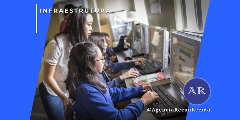 Três jovens estudantes de origem asiática em um laboratório de informática. Uma moça em pé, vestindo camiseta branca, observa atentamente o monitor de outra estudante sentada, que digita em um teclado preto. Ao fundo, uma terceira pessoa também utiliza um computador. No canto superior esquerdo, o selo 'INFRAESTRUTURA' em branco e, no inferior direito, o logotipo da Agência Reconhecida com o @AgenciaReconhecida.
