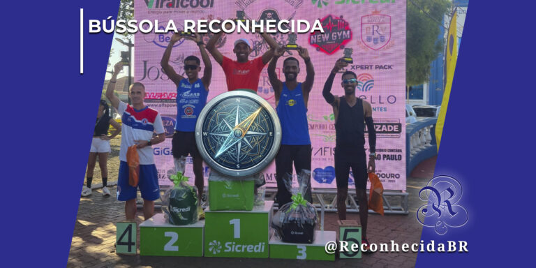 Cinco atletas masculinos de atletismo comemoram em um pódio numerado de um a cinco com a marca Sicredi, segurando pequenos troféus e cestas de premiação em um evento ao ar livre. Ao fundo, um painel rosa exibe logotipos de patrocinadores locais. No centro exato da imagem, há a ilustração sobreposta de uma bússola redonda com detalhes em azul e dourado apontando para o norte. No canto superior esquerdo, aparece o selo BÚSSOLA RECONHECIDA em letras brancas; no inferior direito, o logotipo do portal Reconhecida com o @ReconhecidaBR.