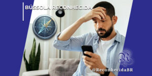 Homem jovem com barba e expressão de preocupação está sentado em um sofá cinza, segurando um smartphone preto com a mão esquerda e levando a mão direita à testa em sinal de desespero ou estresse. No canto superior esquerdo, destaca-se o selo BÚSSOLA RECONHECIDA em letras brancas ao lado da ilustração de uma bússola metálica circular com detalhes dourados e azuis indicando as direções cardeais. No canto inferior direito, aparece o logotipo clássico do portal Reconhecida com o @ReconhecidaBR.