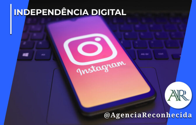 martphone escuro apoiado sobre o teclado de um notebook, exibindo na tela o logotipo clássico do Instagram em destaque com fundo avermelhado e roxo, em um ambiente corporativo escuro. No canto superior esquerdo, lê-se o texto "Independência Digital". No canto inferior direito, o logotipo redondo com as letras "AR" e a assinatura @AgenciaReconhecida.