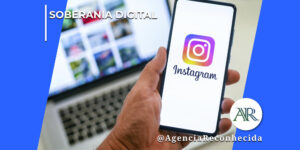Mão masculina segurando um smartphone que exibe o logotipo do Instagram na tela, enquanto ao fundo aparecem a tela de um computador portátil e um teclado em ambiente corporativo. No canto superior esquerdo, lê-se o texto "Soberania Digital". No canto inferior direito, o logotipo redondo com as letras "AR" e a assinatura @AgenciaReconhecida.