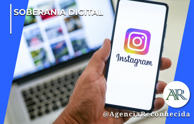 Mão masculina segurando um smartphone que exibe o logotipo do Instagram na tela, enquanto ao fundo aparecem a tela de um computador portátil e um teclado em ambiente corporativo. No canto superior esquerdo, lê-se o texto "Soberania Digital". No canto inferior direito, o logotipo redondo com as letras "AR" e a assinatura @AgenciaReconhecida.